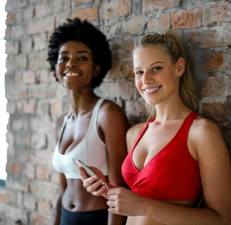 Deux femmes souriantes côte à côte en tenue de sport avec un téléphone à la main devant un mur