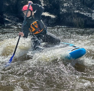 women paddling a Paddleboard over a river wave, wearing helmet, PFD and holding a paddle. 