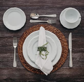 Mesa posta com sousplatt de rattan e jogo de louças de porcelana branca com talheres prateados.