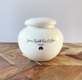 Rounded white ceramic urn with glossy finish, black script text “Sienna Elizabeth Rose O'Shea” and a small rainbow con.