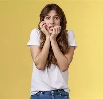 Femme blonde avec visage effrayé bras droit sur joue et doigts main gauche dans la bouche