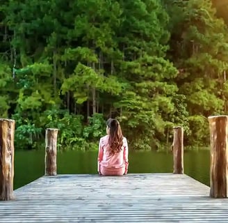Femme de dos cheveux longs habits roses assise sur un ponton au bord d'une rivière bordée d'arbres