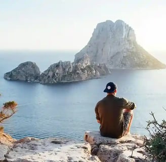 Homme avec 1 casquette visière en AR assis sur 1 rocher face à 1 gros promontoir rocheux dans la mer
