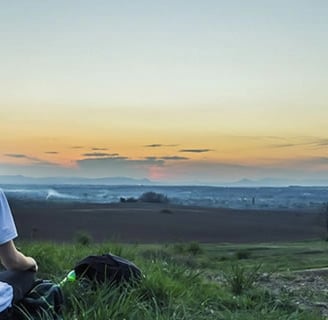 Jeune homme de dos assis dans l'herbe admirant un paysage lointain et un coucher de soleil
