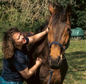 Gemma and a bay horse smiling in the sunshine whilst the horse is getting a massage on their neck.