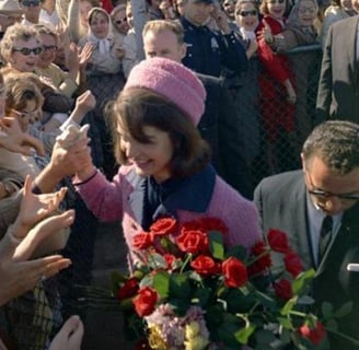 Jackie in a pink coat and hat is surrounded by a crowd of people