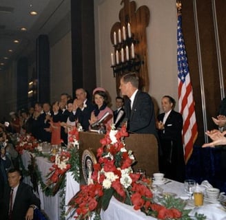 JFK in a suit and tie standing at a podium with a microphone