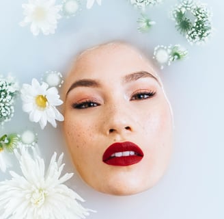 In einer Badewanne mit Milchwasser schaut ein Frauengesicht heraus, um sie herum schwimmen Blumen