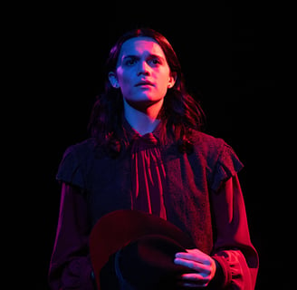 Witch - A distraught actor holding a hat to their chest, washed in red, blue, and purple light