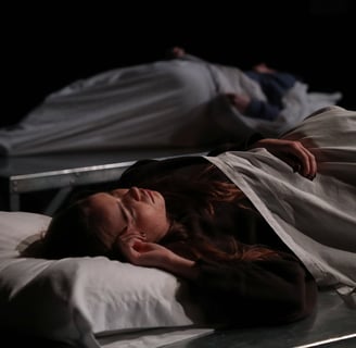 21 North - Two actors laying on beds, one in the foreground and one in the background out of focus.