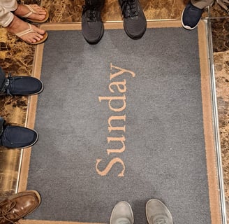 A rug that says "Sunday" with several pairs of feet standing around it