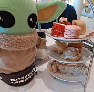 Little Green companion standing beside three tiered plate of mini cakes and tea sandwiches