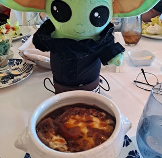 Little green companion wearing a black suit standing in front of bowl of French onion soup 