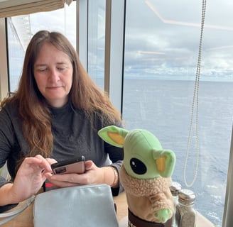 a woman sitting at a table looking at phone with a little green companion