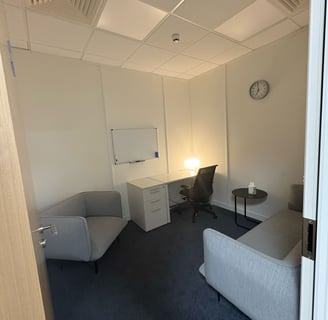 A therapy room, a grey armchair to the left with a sofa to the right, a desk on the back wall