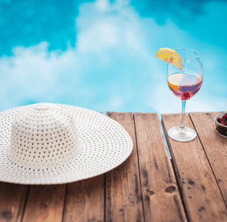 Un verre de rosé avec tranche de citron et un chapeau de plage blanc posés au bord d'une piscine 