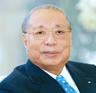 Dr. Daisaku Ikeda looking at the camera wearing golden glasses, a blue suit, and a light blue tie.