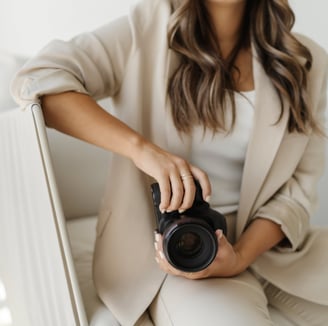 A woman in a beige jacket and white top, sitting on a chair holding a camera.