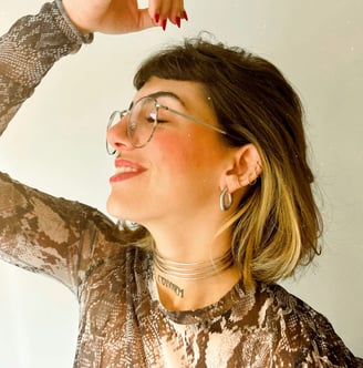 Foto de mulher sorrindo com óculos de grau piercings na orelha e boca, brincos e chocker no pescoço.