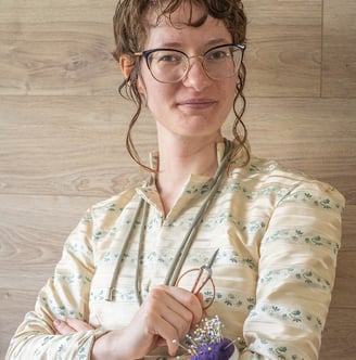 Une personne souriante sur un fond de bois. Elle a des outils de couture en main
