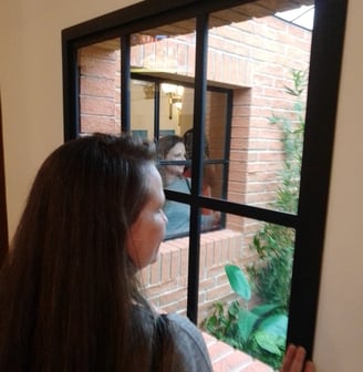 Mulher olha para um jardim pela janela e na lateral esquerda ela se vê refletida.