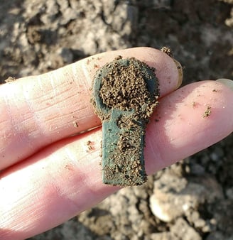 A copper alloy Medieval buckle with green patina, partially covered in soil, held on two fingers.