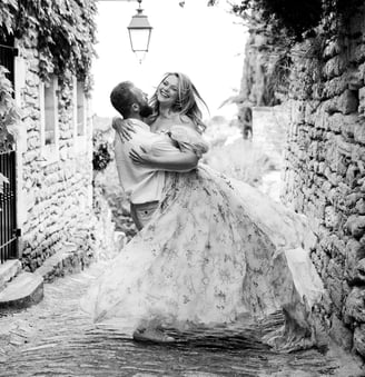 Black and white editorial engagement photo – couple laughing as groom twirls bride in village alley