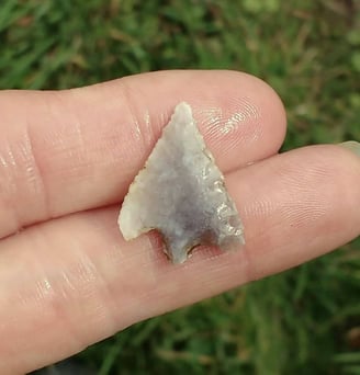 A tiny arrow shaped flint weapon, light grey, held on two fingers.