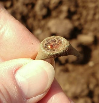 A broken bronze finger ring with green patina and orange inset stone, held between finger and thumb.