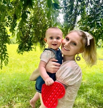 Mother holding baby close under a tree, both smiling at the camera, baby holding heart-shaped toy.
