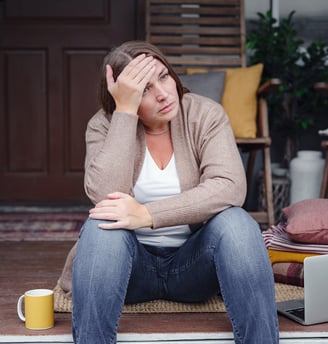 Middle-aged woman sitting on her front porch, hand on forehead, looking tired and reflective. A lapt