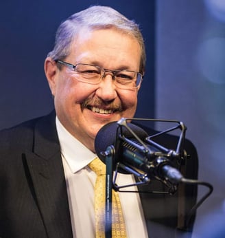 Man in suit and gold tie smiling behind a microphone.