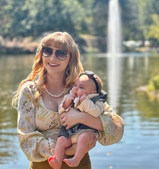 A mother cradling her newborn daughter in front of a lake, with a fountain softly spraying in the background.