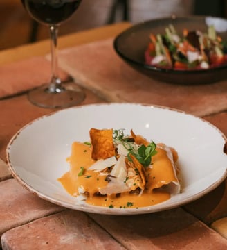 Canelón de temporada con salsa de sobrasada y crujientes, acompañado de una copa de vino tinto en ambiente rústico.