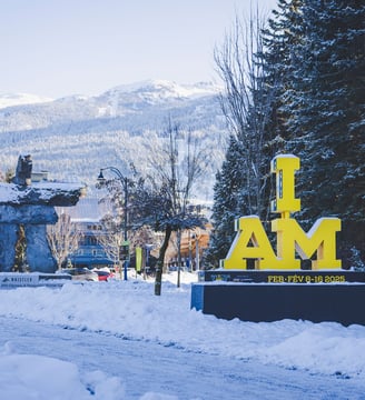 A yellow "I AM" Invictus Games sign in front of a snow-covered Whistler village 