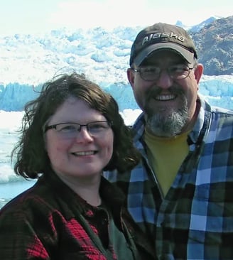 Photo of Mary and her husband Scott- both wearing plaid - with Sawyer Glacier behind them.