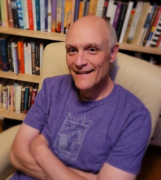 Gregory Spatz sits in an armchair in front of a bookshelf. He wears a purple shirt and smiles.