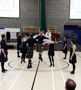 Dancers, dressed mostly in black, dancing a strathspey circle. Judges in the background on a stage.