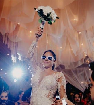 Novia levantando ramo de boda en la mano derecha con lentes de sol con las iniciales de ella y novio