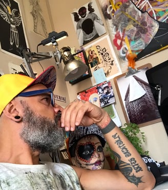 Cenário do ateliê com o artista sentado, refletindo com esboços espalhados pela mesa.