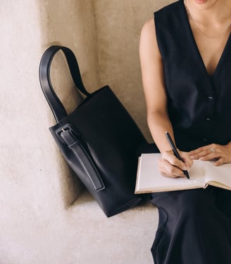 A woman in a sleeveless black dress, sitting and writing in a notebook.