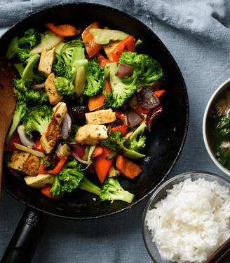 Un wok avec un mélange de légumes et un bol de riz évoquant un repas sain et équilibré asiatique.