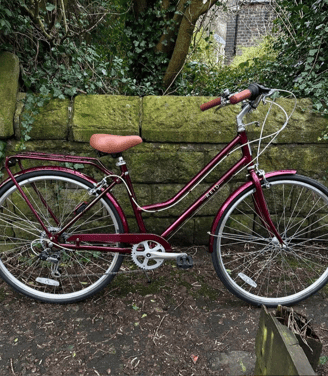 Vélo de ville Reid Classic bordeaux pour femme, avec selle et poignées marron, posé contre un mur de