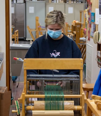 A young woman standing in front of a table loom adjusting your piece.