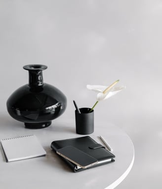 A black vase, pencil cup with a a pen and flower in it, and notebooks on a table