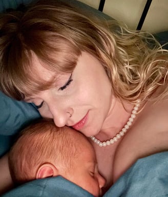 A mother with blonde hair and a pearl necklace peacefully cuddles her sleeping newborn baby, both wrapped in a blue blanket