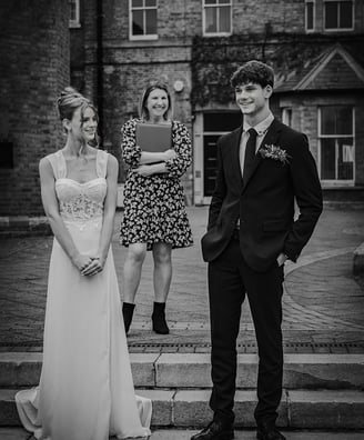A black & white photo of a bride & groom smiling at each other on steps, officiant behind them, in front of a brick building.