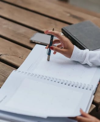 Mão a apontar para calendário mensal num planeador, com caneta e caderno sobre mesa de madeira, repr