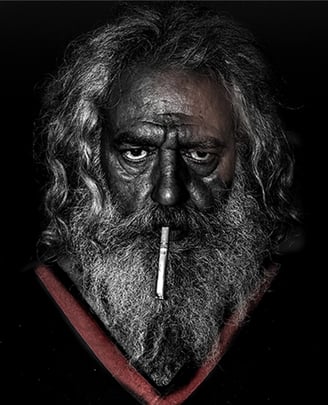 Portrait of an older man with a weathered face and intense gaze, cigarette held between his lips.
