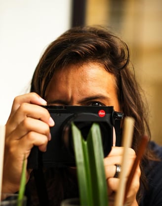 Close‑up of a person with a Leica camera, blurred green leaves in foreground - By ACAT Photos